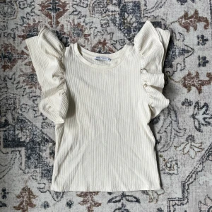 Ribbad volangtopp från Zara - Superfin ribbad topp från Zara med stora volangärmar som ger en cool statement-look. Toppen är ärmlös med volangdetaljer längs axlarna och har en rund halsringning. Perfekt för dig som vill sticka ut lite extra.