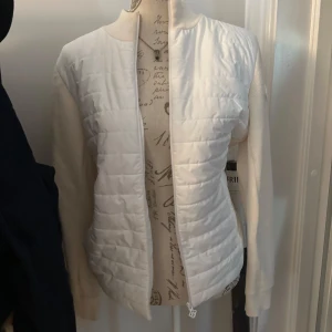 Vit quiltad jacka från J.Lindeberg - Snygg vit jacka från J.Lindeberg med quiltad front och ärmar i stickad struktur. Jackan har dragkedja framtill, ribbad krage och diskret logga på ärmen samt J.Lindeberg-text baktill. Perfekt för dig som gillar stilren och sportig look. Använt 2ggr☺️