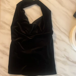 Svart satin bandeau tubtopp - Snygg svart bandeau tubtopp i glansigt satinliknande material. Toppen har en draperad halsringning och är axelbandslös, perfekt för att skapa en trendig look. Passformen är tight och toppen är enkel att styla till olika outfits.