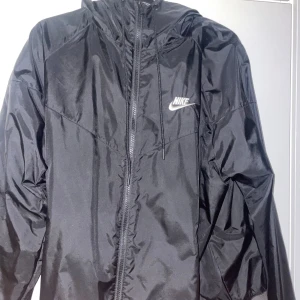 Svart Nike vindjacka med huva M - Svart vindjacka från Nike i storlek M. Jackan har huva med snörning, dragkedja framtill och ribbad nederkant. Tillverkad i lätt och glansig polyester, perfekt för en sportig look. Klassisk Nike-logga på bröstet och enkel, clean design. Dock är jackan lite solbränd.
