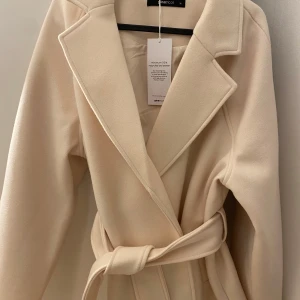 Beige kappa med bälte från Gina Tricot - Snygg beige kappa från Gina Tricot i storlek M. Klassisk modell med bred krage och knytskärp i midjan. Tillverkad av mjuk polyesterblandning, perfekt för dig som vill ha en stilren och enkel look. Passar till många olika outfits!