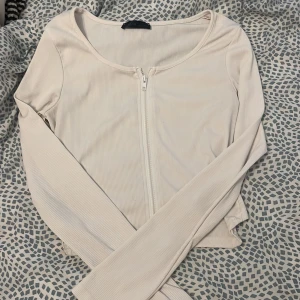 Beige ribbad kofta med dragkedja - Snygg vit ribbad kofta med lång ärm och dragkedja framtill. Modellen är croppad och har en rundad halsringning. Perfekt att slänga över ett linne eller t-shirt för en chill look.