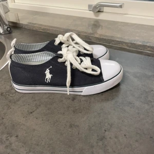 sneakers från Ralph Lauren som NY - Använd 1 gång Snygga mörkblå sneakers från Ralph Lauren med vit logga på sidan, vita snören och tåhätta. Platt sula och rund tå ger en clean och tidlös look. Perfekta för dig som gillar stilrena och sportiga skor.