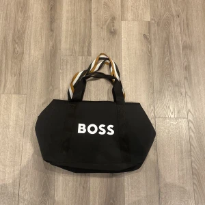 Svart gymväska från BOSS - Stor svart gymväska från BOSS med vita bokstäver framtill. Väskan har breda handtag i svart, vitt och gult med randigt mönster. Tillverkad i slitstark polyester och perfekt för att bära med sig allt man behöver under dagen.
