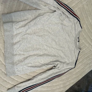 Grå sweatshirt med ränder på ärmarna - Säljer en ljusgrå sweatshirt med rund hals och långa ärmar. Ärmarna har snygga ränder i marinblått, rött och vitt längs sidorna som ger en sportig vibe. Perfekt för chill dagar eller när du vill ha en enkel men stilren look.