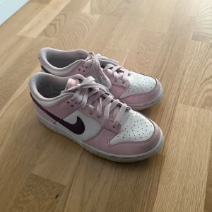 Nike Dunk Low rosa och vita sneakers - Nike Dunk Low sneakers i en snygg kombo av ljusrosa och vitt med mörklila swoosh och broderad Nike-logga på hälen. Skorna har klassisk snörning, perforerad tå och platt sula. Perfekta för dig som vill sticka ut med färg men ändå hålla det stilrent. Äkta 