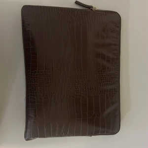 Brun datorväska i skinn med croco-mönster - Snygg brun datorväska från Åhléns i skinn med croco-mönster på ena sidan och slät yta på andra. Datorväskan har dragkedja och ett stort fack med innerficka, perfekt för laptop och dokument. Stilren design med exklusiv känsla.