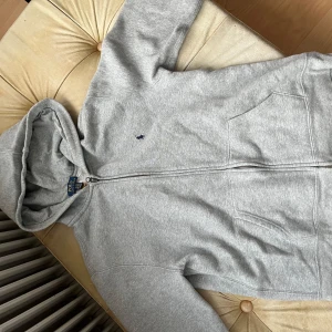 Grå hoodie från Polo Ralph Lauren - Snygg grå hoodie från Polo Ralph Lauren med dragkedja och huva. Klassisk design med broderad logga på bröstet och två stora fickor framtill. Tillverkad i mjuk bomullsmix som är perfekt för chill dagar. Passar dig som gillar enkel och stilren streetwear.
