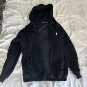 Svart hoodie från Polo Ralph Lauren - Svart hoodie från Polo Ralph Lauren med dragkedja och huva. Klassisk design med vit broderad logga på bröstet, två fickor framtill och ribbade muddar. Perfekt för en chill och stilren look. Storlek L men sitter som S/M.