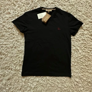 Burberry- svart slim fit tshirt  - Svart t-shirt från Burberry i mjuk bomull med klassisk rund hals och korta ärmar. Diskret broderad Burberry-logga i vinrött på bröstet och ikoniskt rutigt band på insidan av kragen. Perfekt för en clean och stilren look. Den är helt ny och oanvänd med alla lapparna kvar. Den har en helt sjuk fit som får en att se mycket större ut (bra för de som gymmar). 