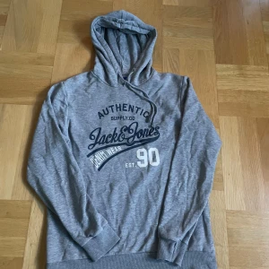 Grå hoodie från Jack & Jones - Snygg grå hoodie från Jack & Jones med tryck på bröstet och klassisk huva med snörning. Mjuk bomullskänsla och ribbade muddar vid ärmslut och nederkant. Perfekt för chill dagar eller när du vill ha en avslappnad stil.
