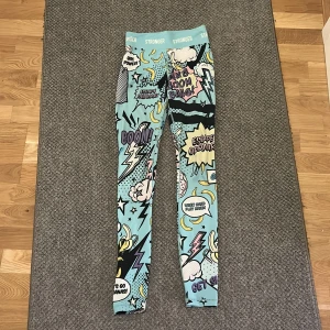 Stronger leggings med pop art-mönster - Färgglada leggings från Stronger med pop art-inspirerat tryck i blått, gult, rosa och svart. Hög midja och tight passform, perfekta för träning eller när du vill sticka ut. Tillverkade i stretchigt syntetmaterial för maximal komfort.