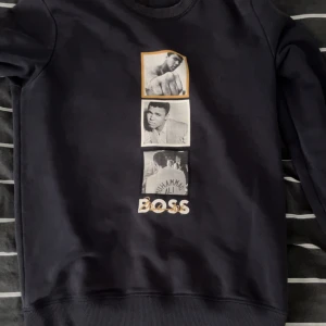 Svart BOSS x Muhammad Ali sweatshirt - Svart sweatshirt från BOSS x Muhammad Ali med tryck av tre svartvita bilder och texten 'BOSS' framtill. Tröjan har rund hals, långa ärmar och är gjord i mjuk bomullsmix. Perfekt för dig som gillar sportiga och stilrena plagg med unik design.