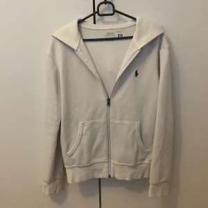 Vit hoodie från Polo Ralph Lauren XS - Snygg vit hoodie från Polo Ralph Lauren i storlek XS. Klassisk design med dragkedja, huva och två fickor framtill. Broderad logga på bröstet. Perfekt för en chill och stilren look. Den har inga defekter alls den är helt ren och ny. Finns inga snören till tröjan !