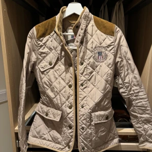 Beige quiltad jacka från Lexington XS -  beige jacka från Lexington med bruna axelpartier i mocka-look. Jackan har broderad logga på bröstet, tryckknappar, fickor med lock och markerad midja. Perfekta jackan till hösten. Fint skick använt ett fåtal gånger 