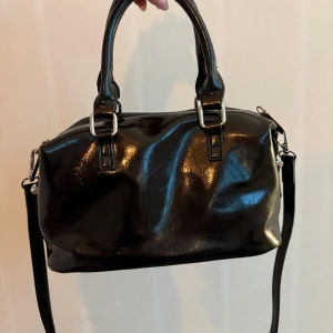 Svart handväska i glansigt skinn - Snygg svart handväska i glansigt skinn med både handtag och avtagbar axelrem. Väskan har silverfärgade metalldetaljer och dragkedja upptill. Stilren och enkel design som passar till många outfits.