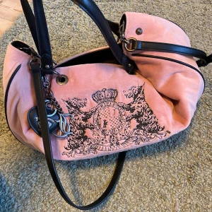 Juicy Couture vintage double handle bag - Säljer en rosa axelväska från Juicy Couture i sammetstyg med svarta detaljer och handtag. Väskan har broderat Juicy Couture-emblem framtill, snygga metalldetaljer och innerfoder med logotypmönster.    Höjd: 27 cm, Längd: 36 cm, Vidd: 11 cm, Hantag: 16.5 cm. Använd 2-3 ggr, så praktiskt taget helt ny!