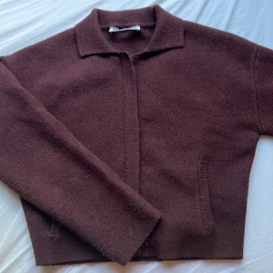 Vinröd stickad cardigan jacka från Zara - Mysig vinröd stickad cardigan jacka från Zara med klassisk krage och dragkedja framtill. Jackan har två fickor och en rak, något croppad passform. Perfekt för lager-på-lager och chill vibes under höst och vinter.