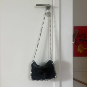 Svart axelväska med kedja från Tiamo - Cool svart axelväska från Tiamo i skinn med silverfärgad kedja som axelrem och snygga detaljer runt kanterna. Väskan har magnetlås och flera fack inuti, perfekt för att hålla ordning på dina grejer. Stilren och edgy design som lyfter vilken outfit som helst.🩷