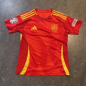 Spaniens landslagströja Adidas röd - Säljer en röd Spanien fotbollströja från Adidas med gula detaljer och tryck nummer 19 samt Lamine Yamal på ryggen. Tröjan har korta ärmar, broderat landslagsmärke på bröstet och klassiska Adidas-ränder på axlarna. Perfekt för dig som vill visa ditt stöd för Spanien!
