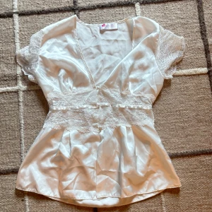 Vit sidenblus med spets från Bondy - Supersöt vit blus i glansigt sidenmaterial från Bondy storlek S, med v-ringad hals och korta spetsärmar. Dekorerad med bred spets och pärlknappar över midjan för extra romantisk vibe. Säljer då jag har aldrig fått chansen att använda den :)