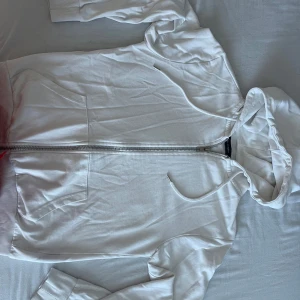 Vit hoodie från The Black Label Collection - Snygg vit hoodie med dragkedja från The Black Label Collection. Tröjan har huva med snören, ribbade muddar och broderad logga på ärmen. Perfekt för en clean och stilren look. Gjord i mjuk bomull för extra komfort.