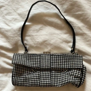 Svartvit rutig axelväska - Snygg axelväska i svartvitt mönster, Väskan har en smal svart axelrem men är lite sliten som syns på sista bilden. Köpt second hand i England så vet ej märket