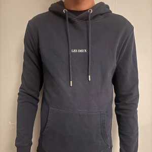 Mörkblå hoodie från Les Deux - Snygg mörkblå hoodie från Les Deux med vit logga på bröstet. Tröjan har huva med snörning, magficka och långa ärmar. Tillverkad i mjuk bomull som är skön att ha på sig. Perfekt för chill dagar och enkel att matcha med jeans eller joggers.