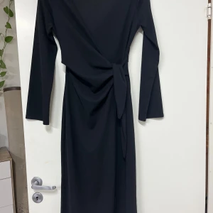Svart omlottklänning från H&M - Snygg svart långklänning från H&M med omlott-design och knyt i midjan. Klänningen har långa ärmar och en stilren, elegant look. Materialet är mjukt och följsamt polyester som ger en skön passform. Perfekt för dig som gillar minimalistisk stil med en twist.