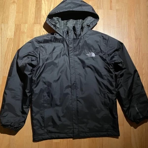 The North Face vindjacka - Svart vindjacka från The North Face med huva och dragkedja framtill. Jackan har en stilren look med logga på bröstet och är tillverkad i ett lätt fodrat, vattenavvisande material. Perfekt för blåsiga dagar och enkel att matcha med allt.