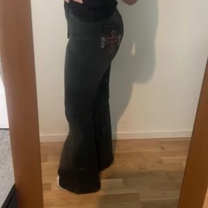 Svarta bootcut byxor med broderad ficka - Säljer ett par svarta bootcut byxor med snygg broderad ficka bak, ett rött kors och vita detaljer. Byxorna har en klassisk bootcut passform som sitter tajt upptill och släpper ut vid benen. Helt oanvända men för högmidjade för min smak.