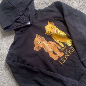Svart Lion King hoodie från Disney - Svart hoodie från Disney med tryck av Simba och Nala från Lejonkungen på framsidan. Tröjan har huva och långa ärmar, perfekt för dig som älskar klassiska Disneyfilmer. Mjuk insida och skön passform.