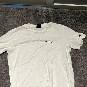 Vit Champion t-shirt med logga - Vit t-shirt från Champion med klassisk logga broderad på bröstet och en liten logga på ärmen. Enkel och stilren design, perfekt till jeans eller shorts. Skön passform och mjukt material.