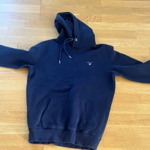 Marinblå hoodie från GANT med huva strl S  - Säljer en marinblå hoodie från GANT i storlek S. Tröjan har broderad logga på bröstet, stor känguruficka och justerbar huva med snörning. Mjuk bomullskänsla och ribbade muddar vid ärmslut och nederkant. 