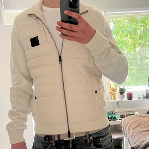 Vit cardigan från BOSS - Snygg vit cardigan från BOSS med quiltad framsida och ribbade muddar. Jackan har dragkedja hela vägen, två fickor med knapp och en svart BOSS-logga på bröstet. Ärmar och baksida är i mjukt tyg för extra komfort. Perfekt för dig som gillar stilren och clean look. Om ni har några funderingar är det bara att fråga, MVH