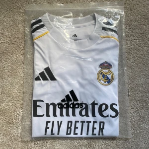 Real Madrid vit matchtröja Adidas - Säljer en officiell Real Madrid fotbollströja från Adidas i vit med svarta detaljer och gula linjer. Tröjan har korta ärmar, klubbmärke på bröstet och Emirates Fly Better-tryck framtill. Tillverkad i lätt funktionsmaterial för bästa komfort på planen.