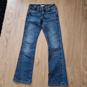 Blå bootcut jeans från Gina Tricot - Snygga blå jeans från Gina Tricot i storlek 164. Modellen har bootcut-ben och klassisk femficksdesign med coola ficklock och silverfärgade knappar bak. Jeansen har normal passform och är tillverkade i mjukt denimtyg med slitningar för en trendig look.