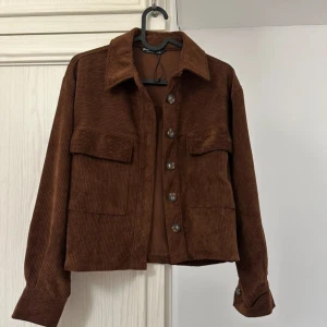Brun manchester overshirt - Snygg brun overshirt i manchester med stora fickor framtill och knappar hela vägen. Jackan har klassisk krage och långa ärmar med knapp vid ärmslut. Perfekt att slänga över en hoodie eller t-shirt för en chill look.