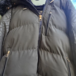 Svart pufferjacka Glorious Gangsta - Snygg svart pufferjacka från Glorious Gangsta med gulddetaljer på dragkedjorna, mönstrade ärmar och stor huva med fejkpäls. Insidan är fodrad med mjuk sammet för extra komfort. Jackan har flera fickor med dragkedja och en patch på ärmen. Knappt använd så väldigt fint skick!