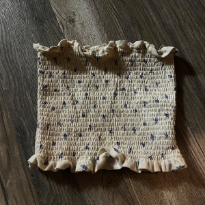 Blommig bandeau tubtopp med volang från H&M - Söt bandeau tubtopp i ljusbeige med små blå blommor och volangdetaljer upptill och nedtill. Den är även väldigt stretchig 