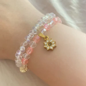 Två pärlarmband med rosa och guld - Säljer två söta pärlarmband, ett med transparenta pärlor och ett med rosa och klara pärlor. Det rosa har en guldig blomberlock med glittriga stenar och små guldplattor. Perfekt för dig som gillar att mixa och matcha smycken med lite extra bling.