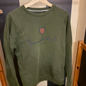 Olivgrön sweatshirt från Bondelid - Olivgrön sweatshirt från Bondelid med tryckt logga och sköld på bröstet. Tröjan har rund hals, långa ärmar och är tillverkad i mjuk bomull. Perfekt för en chill och avslappnad stil.