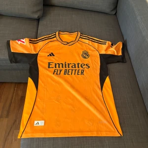 Real Madrid orange matchtröja Adidas - Säljer en snygg orange Real Madrid fotbollströja från Adidas med svarta detaljer och klubbmärke på bröstet. Tröjan har korta ärmar, rund hals och tryckta sponsorer. Perfekt för dig som vill sticka ut på planen eller heja från läktaren.
