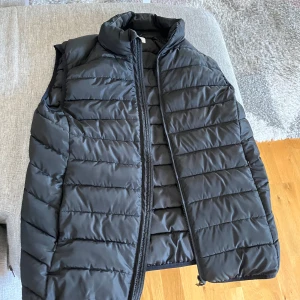 Svart quiltad dunväst med dragkedja - Säljer en svart dunväst med quiltad design och hög krage. Västen har en hel dragkedja framtill och är lätt vadderad, perfekt att slänga över en hoodie eller tröja. Enkel och stilren look som funkar till det mesta.