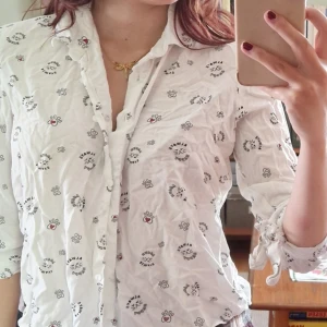 Vit skjorta med tryck från FB Sister - Säljer en vit skjorta från FB Sister i storlek XS med söta små tryck av tassar och hjärtan. Skjortan har långa ärmar med knyt vid ärmsluten och knappar framtill. Perfekt för dig som gillar unika detaljer och en lekfull stil.