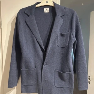 Cardigan från John Henric - Snygg blå cardigan från John Henric med klassisk krage, tre fickor framtill och knäppning med en knapp. Jackan är stickad och har lång ärm, perfekt för dig som gillar stilren och enkel design. Sparsamt använd. Passar både S och XS.