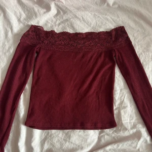 Vinröd offshoulder topp med spets - Vinröd ribbad offshoulder topp med lång ärm och bred spetskant upptill. Snygg och figurnära modell som ger en cool och romantisk vibe. Perfekt att styla med jeans eller kjol för en trendig look.