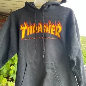 Svart Thrasher hoodie med flames - Svart hoodie från Thrasher med ikoniskt flames-tryck i gult och rött på bröstet. Klassisk känguruficka, huva med snörning och mjukt material. Perfekt för dig som gillar streetwear och skatekultur.