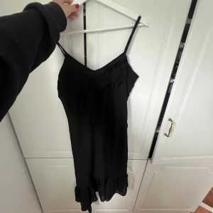 Svart långklänning från SusyMix - Supersnygg svart långklänning från SusyMix med smala axelband, v-ringning och volangdetaljer både upptill och nertill. Klänningen har en flowy passform och är tillverkad i ett lätt, glansigt material som känns lyxigt.