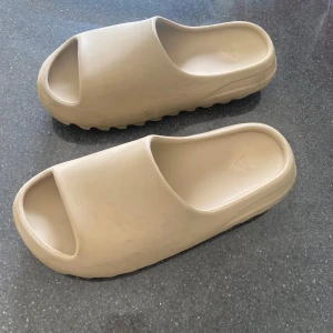Beige Yeezy Slide från Adidas - Säljer ett par Yeezy Slide från Adidas i beige färg. Slidesen har en chunky, modern design med öppen tå och platt sula. Tillverkade i syntetmaterial som är både lätta och bekväma. Perfekta för chill dagar eller när du vill ha en clean look. Storlek 10 US eller 43 EU låda ingår.